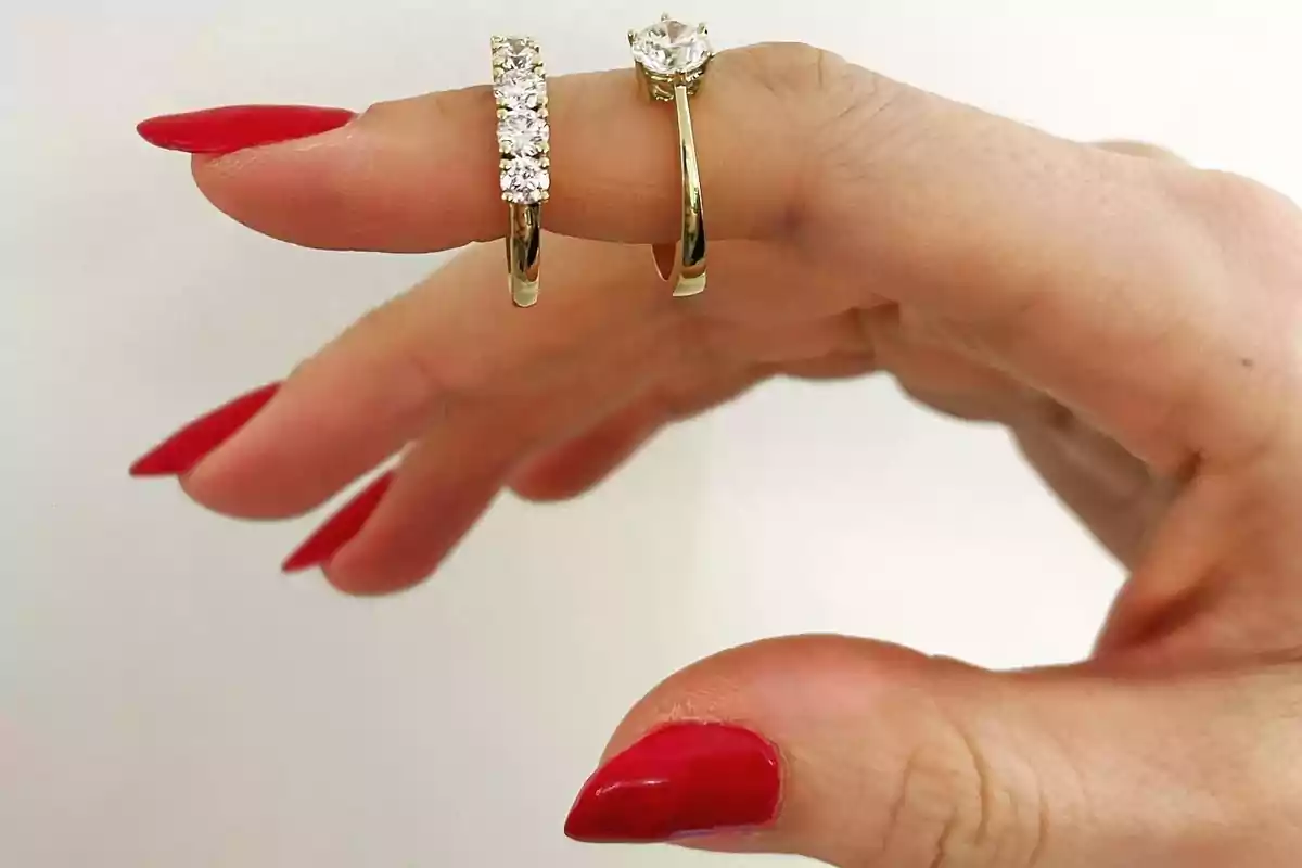 A hand with red nails holds two gold rings with diamonds.