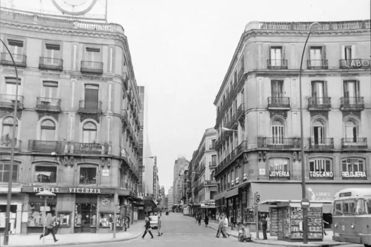 Una calle urbana con edificios antiguos y tiendas en las plantas bajas, personas caminando y un autobús en la esquina.