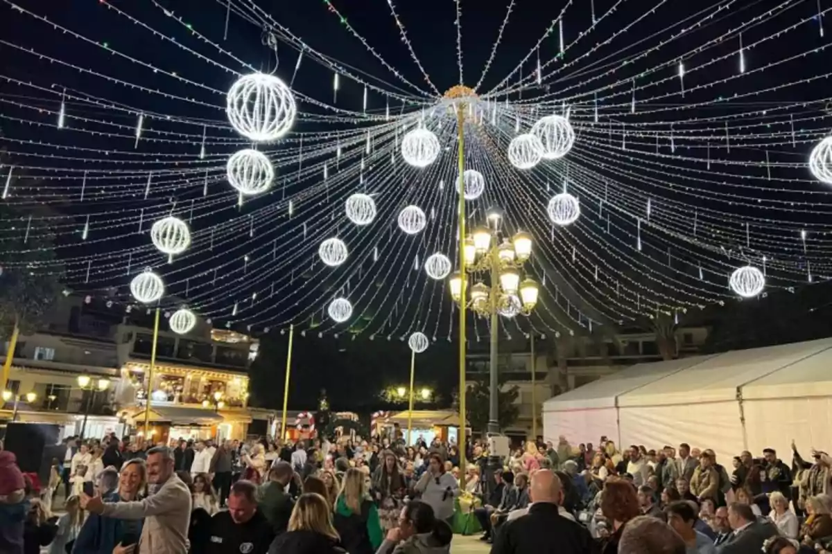 Una multitud disfruta de un evento nocturno al aire libre bajo un techo de luces decorativas en forma de esferas brillantes.