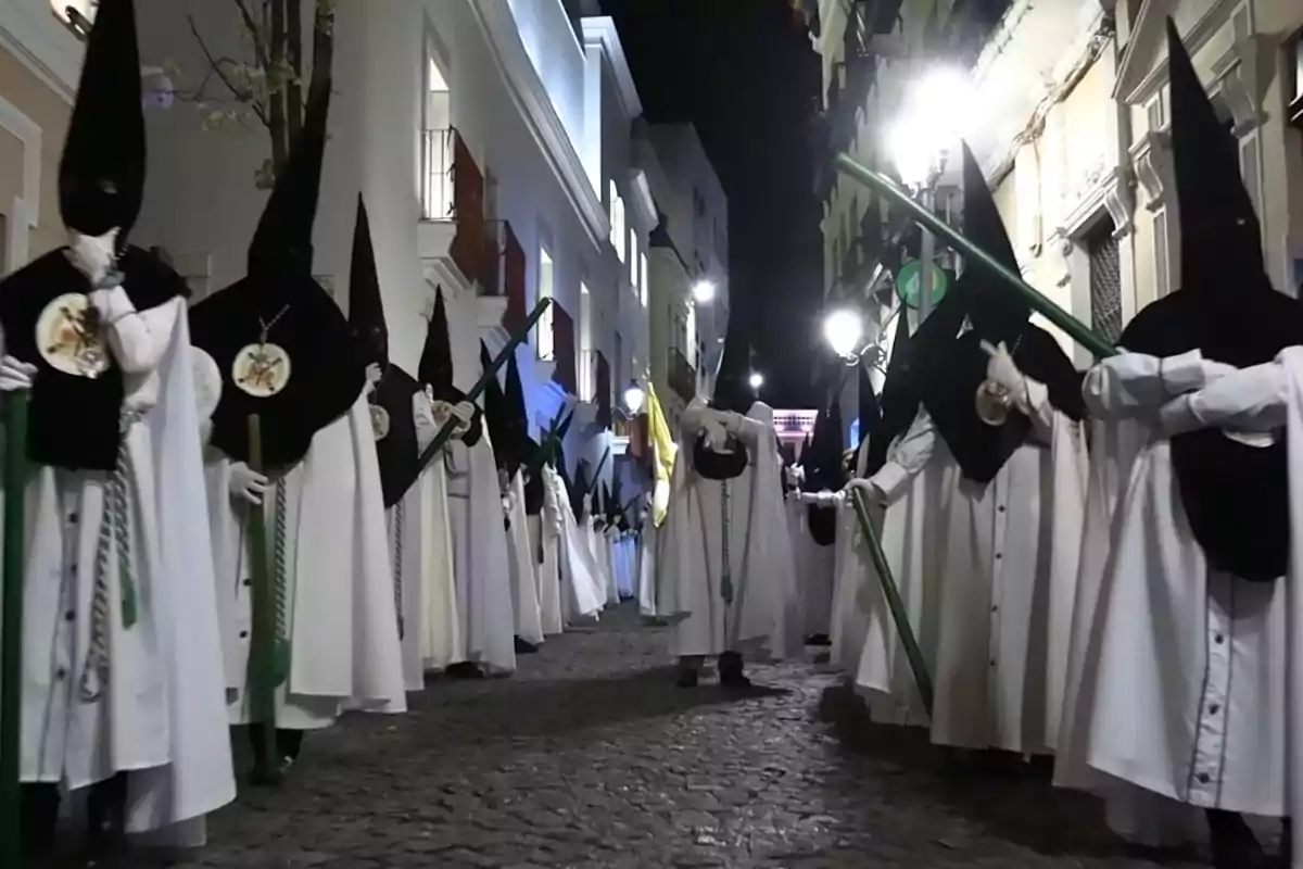 Un grupo de personas vestidas con túnicas blancas y capirotes negros camina por una calle empedrada durante la noche. Un grupo de personas vestidas con túnicas blancas y capirotes negros camina por una calle empedrada durante la noche.