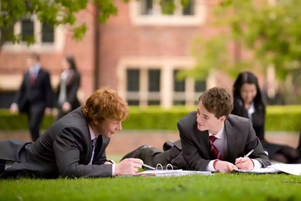 Dos estudiantes con traje y corbata están acostados en el césped mientras estudian y conversan en un entorno universitario al aire libre.