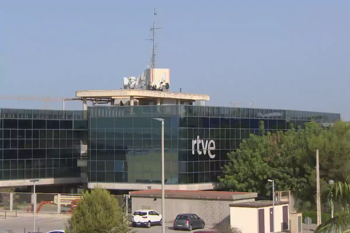 Edificio moderno de cristal con el logo de rtve rodeado de árboles y algunos coches estacionados en primer plano Edificio moderno de cristal con el logo de rtve rodeado de árboles y algunos coches estacionados en primer plano