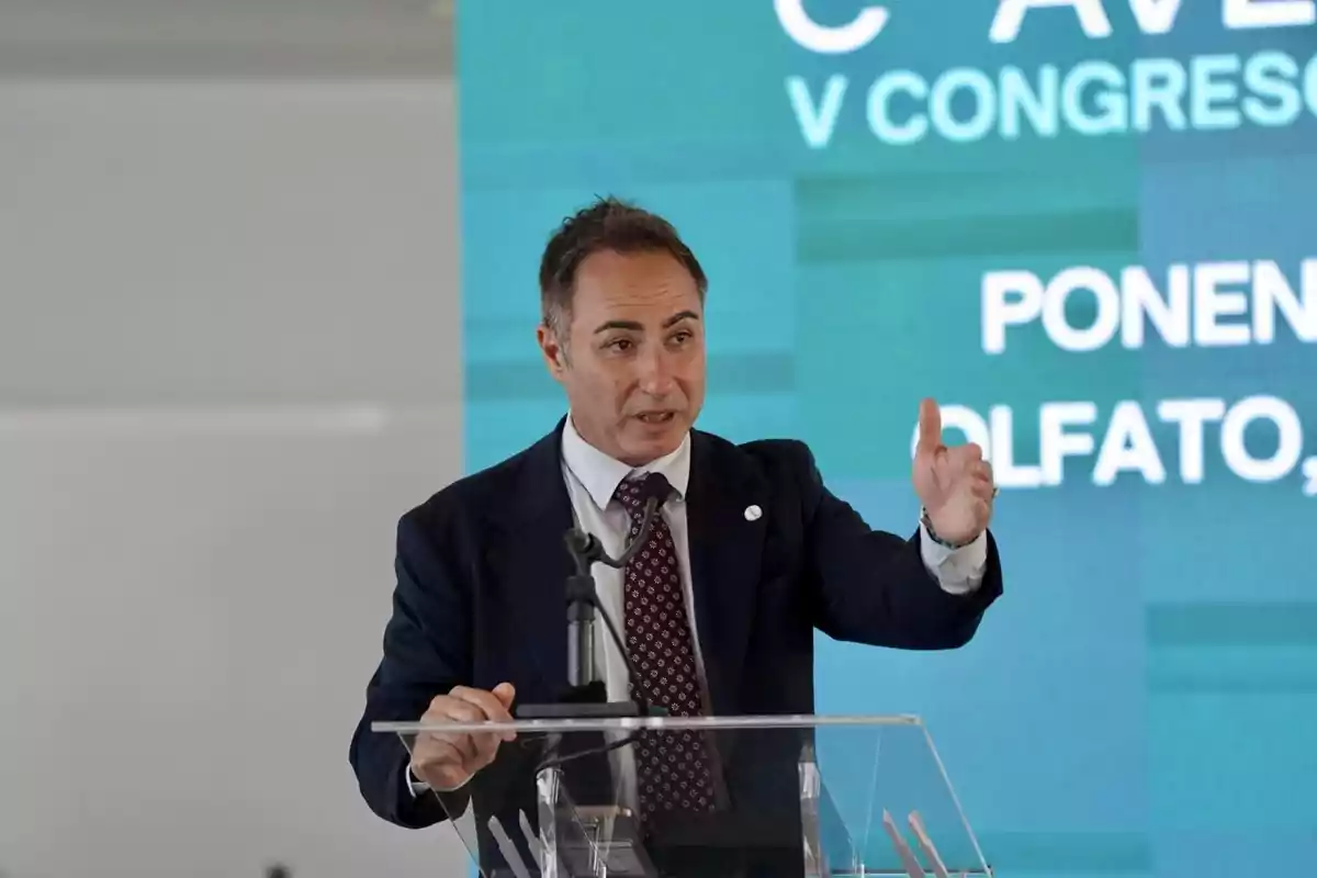 Hombre de traje hablando en un podio con micrófono y fondo azul con texto de congreso