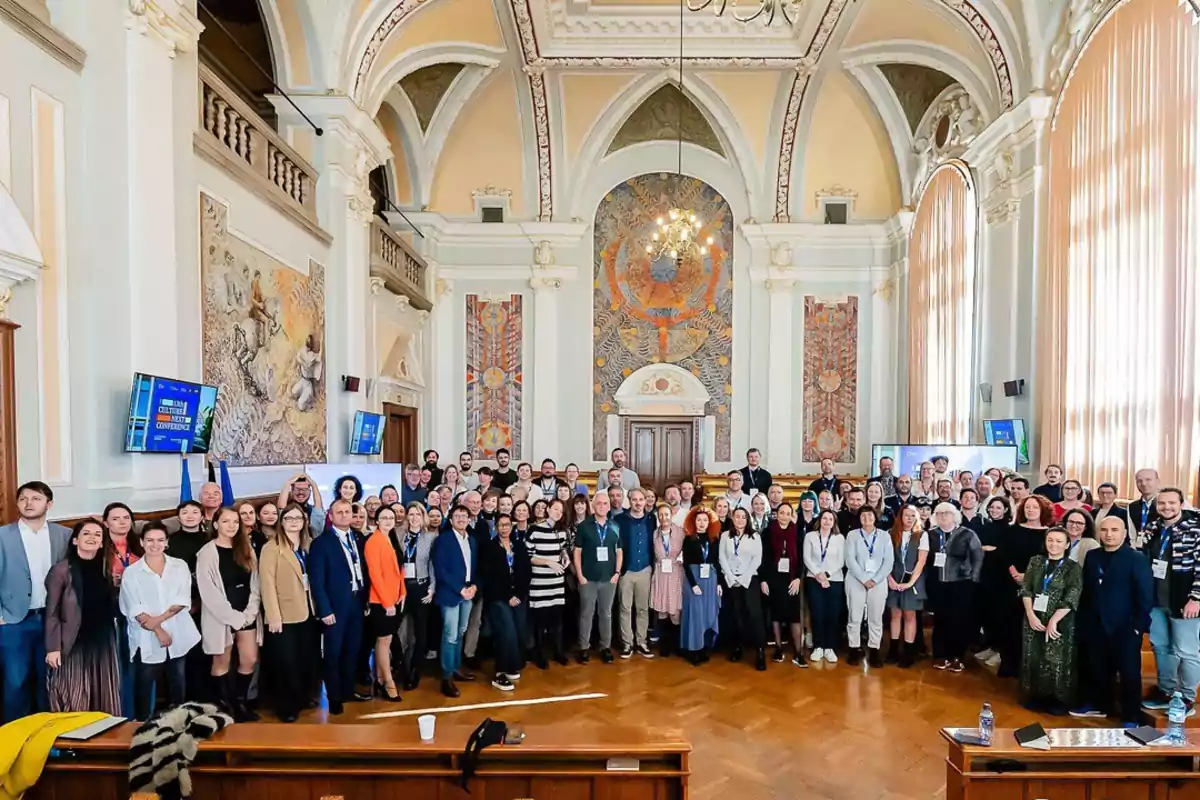 Un gran grupo de personas posando para una foto en el interior de un elegante salón con techos altos, vitrales y murales decorativos