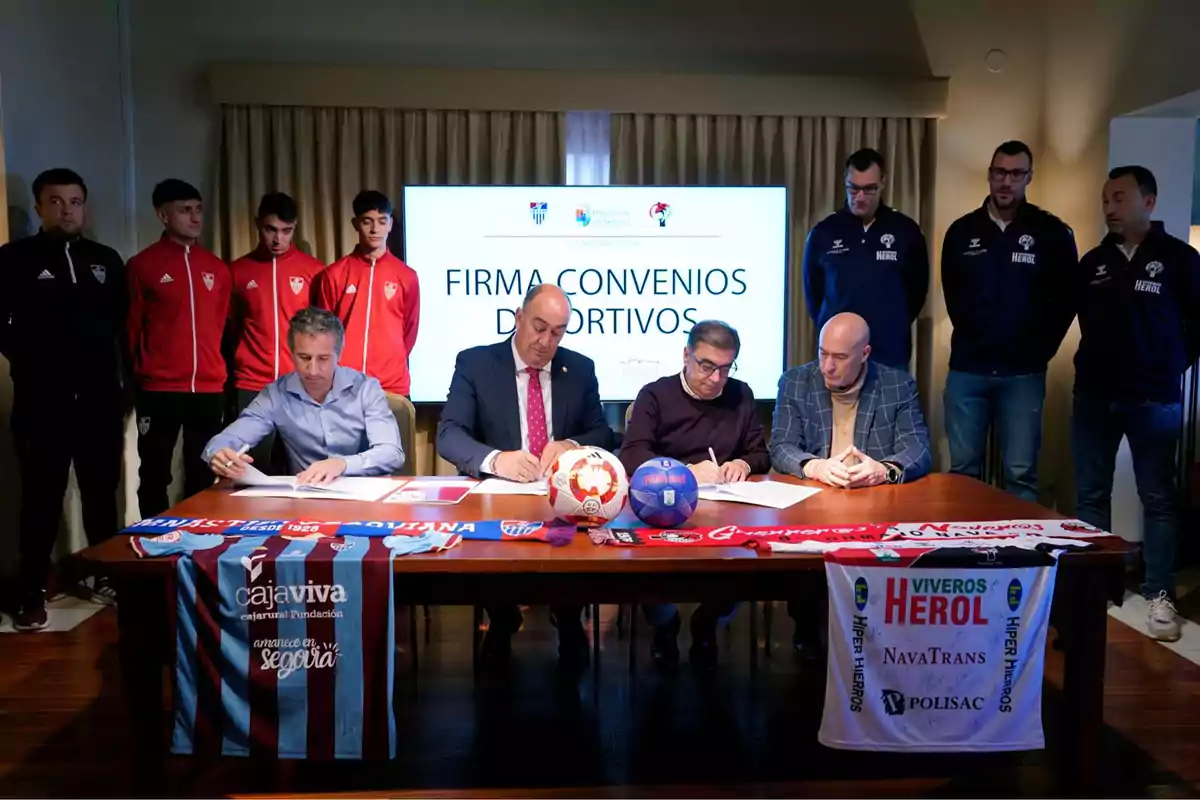 Un grupo de personas firma convenios deportivos en una mesa con bufandas y balones, mientras otros observan de pie.