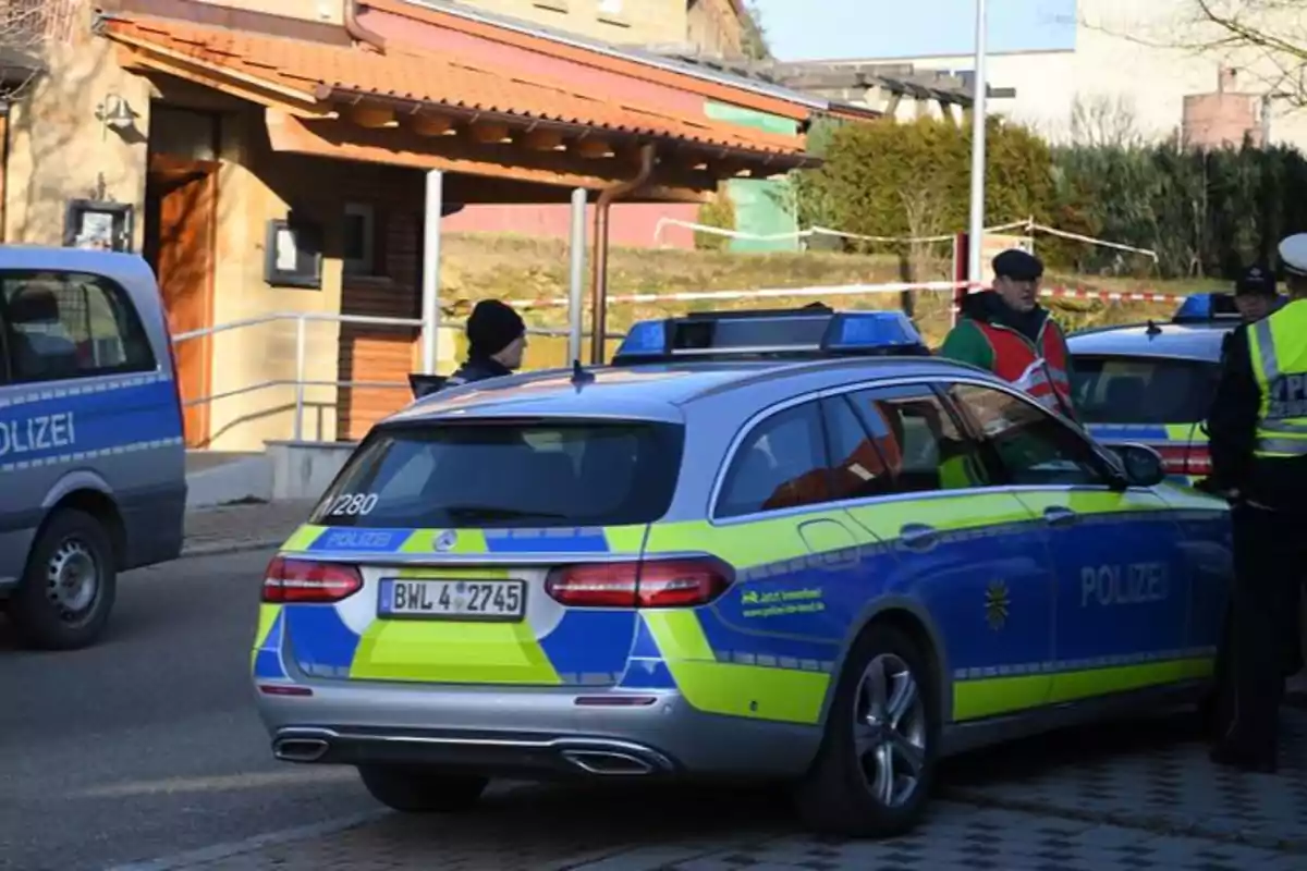 Una escena muestra varios vehículos de la policía y oficiales en una calle frente a un edificio con techo de tejas.