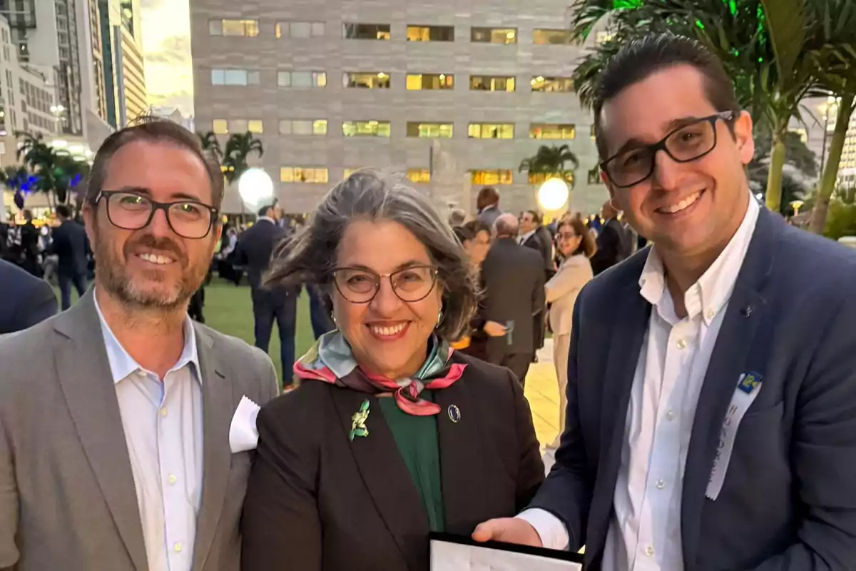 Tres personas sonríen en un evento al aire libre con un edificio de fondo.