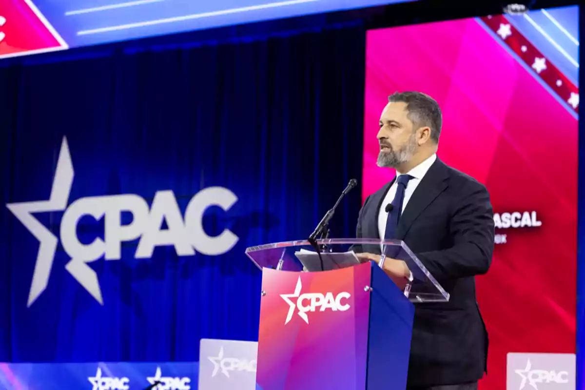 A man in a suit speaks at a podium during a CPAC event with a blue and red background.