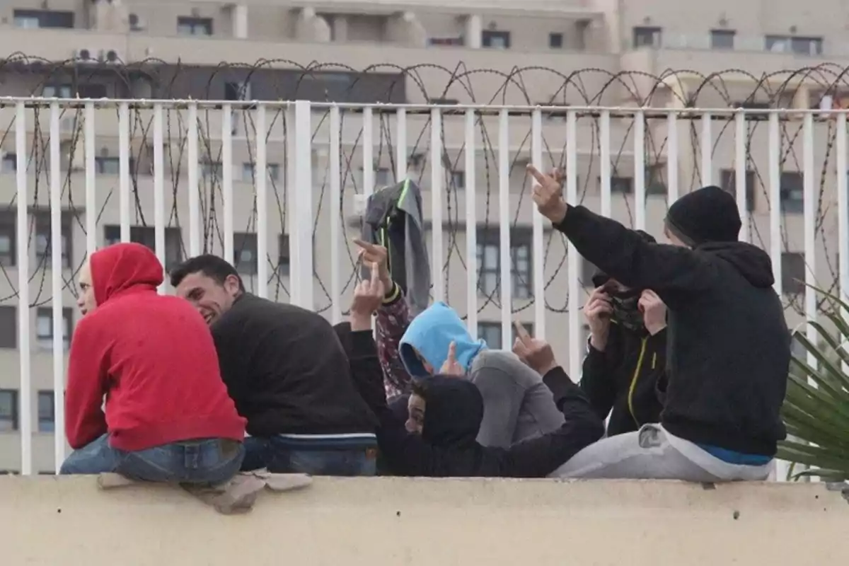 Un grupo de jóvenes con sudaderas y capuchas se encuentra sentado en una estructura elevada, algunos de ellos haciendo gestos con las manos, con un fondo de edificios y una cerca con alambre de púas.