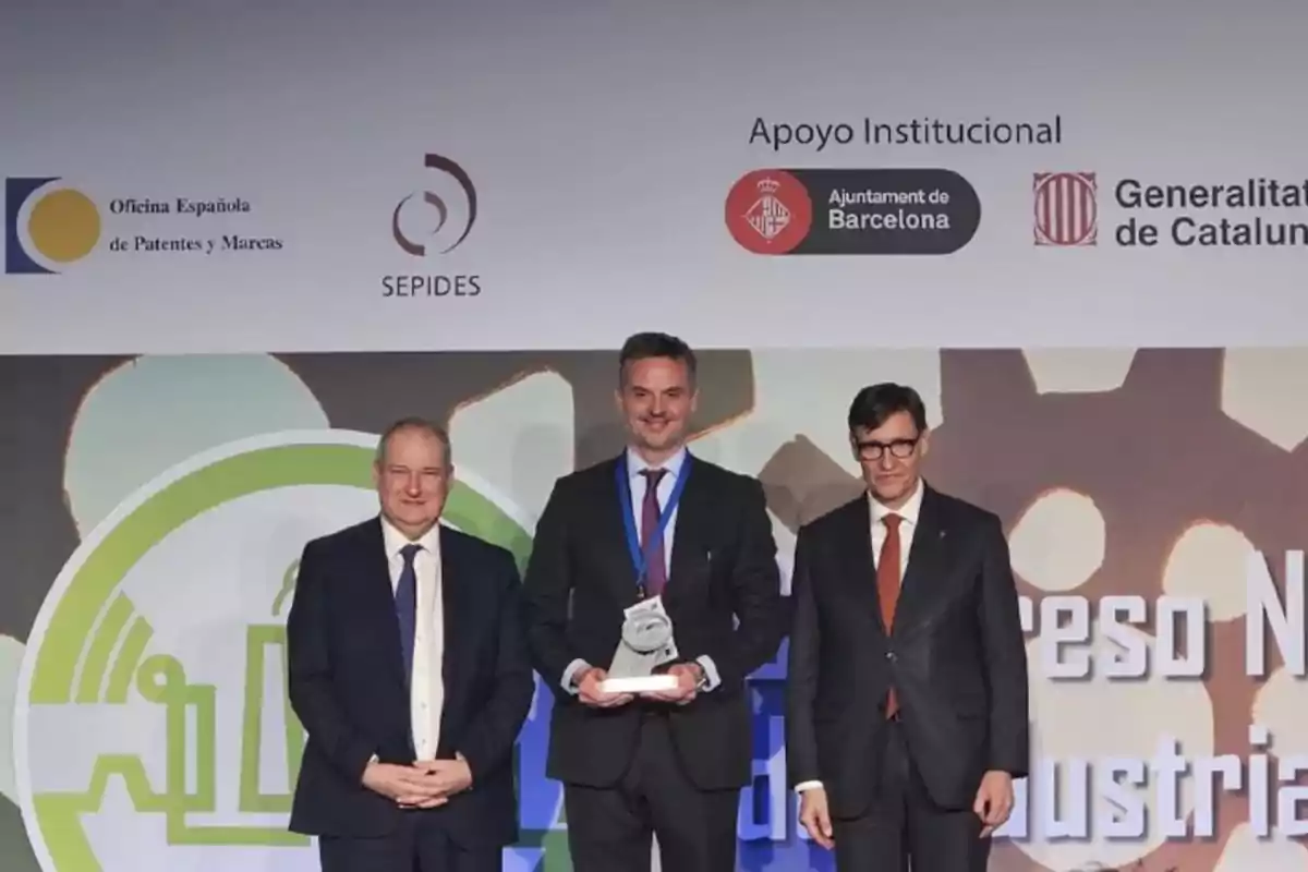 Tres hombres en traje posan en un evento de premiación con logotipos de la Oficina Española de Patentes y Marcas, SEPIDES, Ayuntamiento de Barcelona y Generalitat de Catalunya en el fondo.