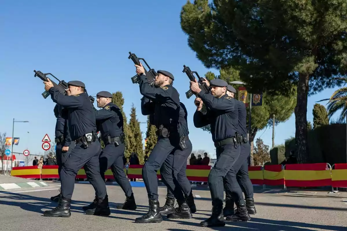 Group of police officers in blue uniform marching with weapons in parade position on a street with flags and trees in the background.