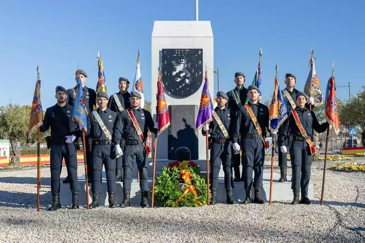 A group of uniformed people hold flags in front of a monument with a wreath at its base.