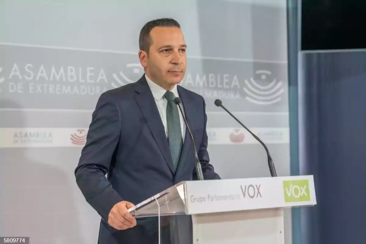 Un hombre de traje oscuro y corbata verde está de pie en un podio con micrófonos en la Asamblea de Extremadura.