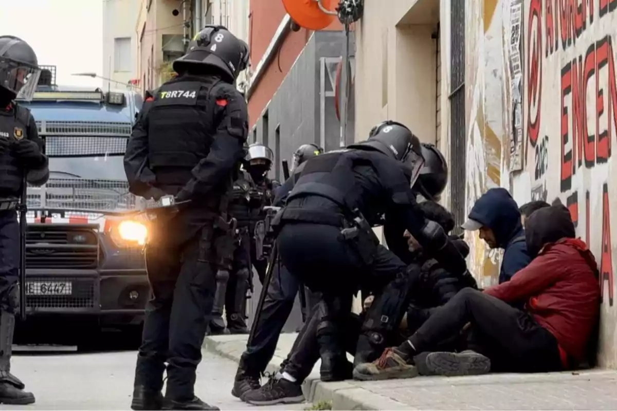 Un grupo de policías antidisturbios interactúa con varias personas sentadas en la acera junto a un vehículo policial en una calle urbana. Un grupo de policías antidisturbios interactúa con varias personas sentadas en la acera junto a un vehículo policial en una calle urbana.