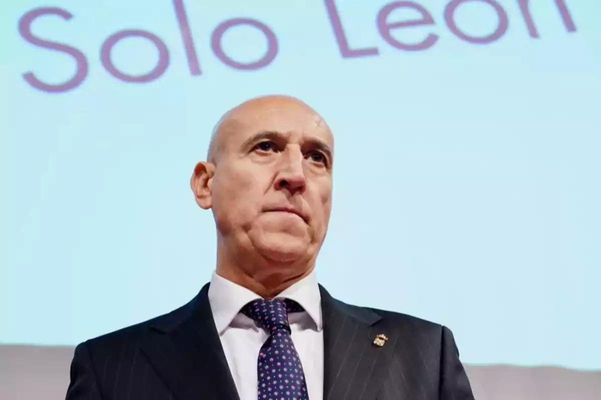Hombre calvo con traje oscuro y corbata de puntos de pie frente a una pantalla con letras desenfocadas al fondo Hombre calvo con traje oscuro y corbata de puntos de pie frente a una pantalla con letras desenfocadas al fondo