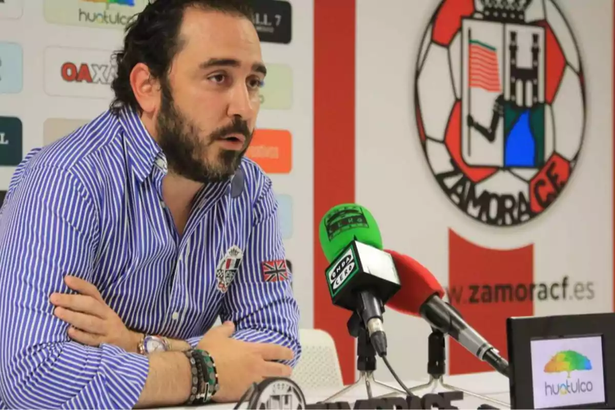 Hombre con barba y camisa de rayas azules hablando en una rueda de prensa con micrófonos frente a él y el escudo del Zamora CF en el fondo