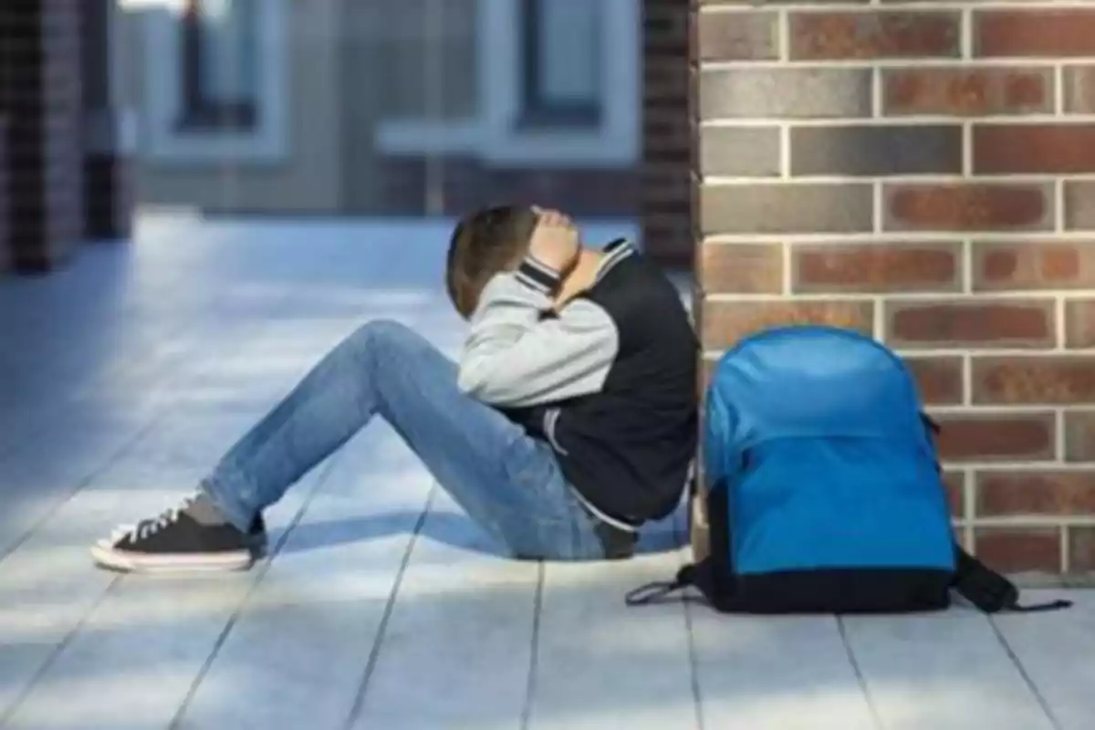 Un niño sentado en el suelo con la cabeza entre las manos junto a una mochila azul.