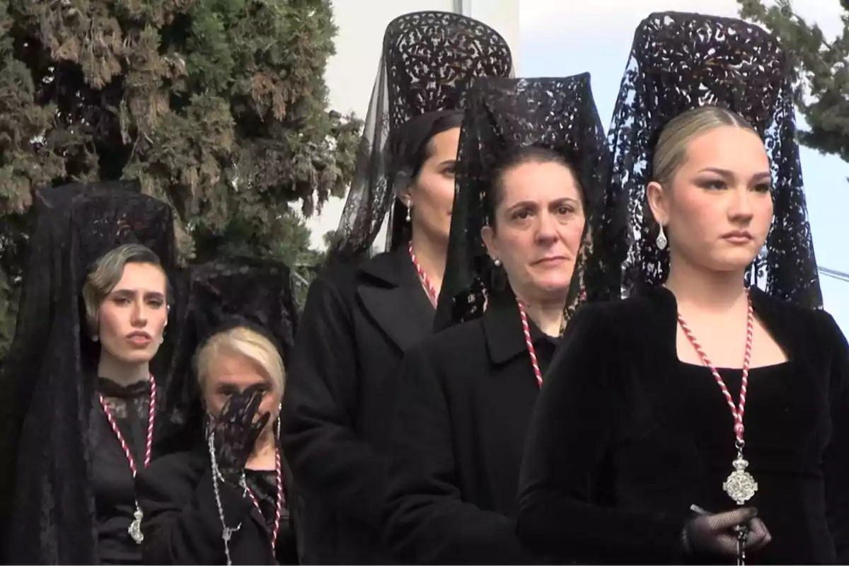 Un grupo de mujeres vestidas de negro con mantillas y collares, participando en una procesión religiosa al aire libre. Un grupo de mujeres vestidas de negro con mantillas y collares, participando en una procesión religiosa al aire libre.