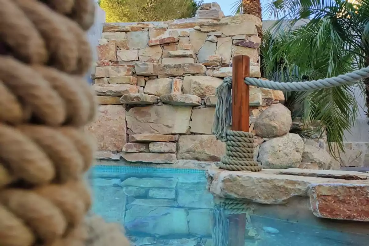 Una piscina rodeada de piedras y vegetación con una cuerda gruesa enrollada en un poste de madera.