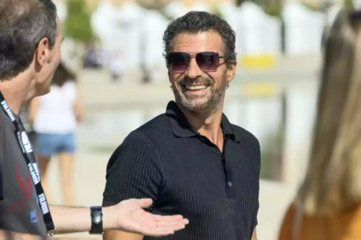 Un hombre con gafas de sol y barba sonríe mientras conversa con otras personas al aire libre.