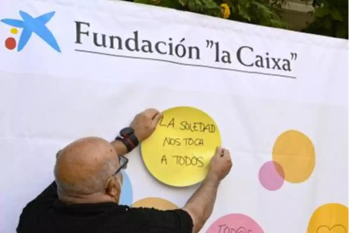 Un hombre coloca un cartel amarillo con el mensaje "La soledad nos toca a todos" en un mural de la Fundación "la Caixa".