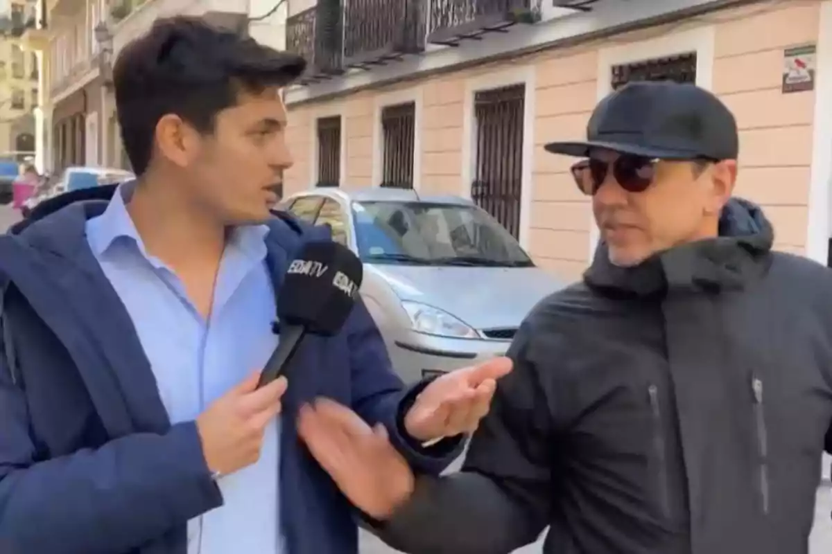 A reporter holds a microphone while interviewing a man with sunglasses and a cap on an urban street.