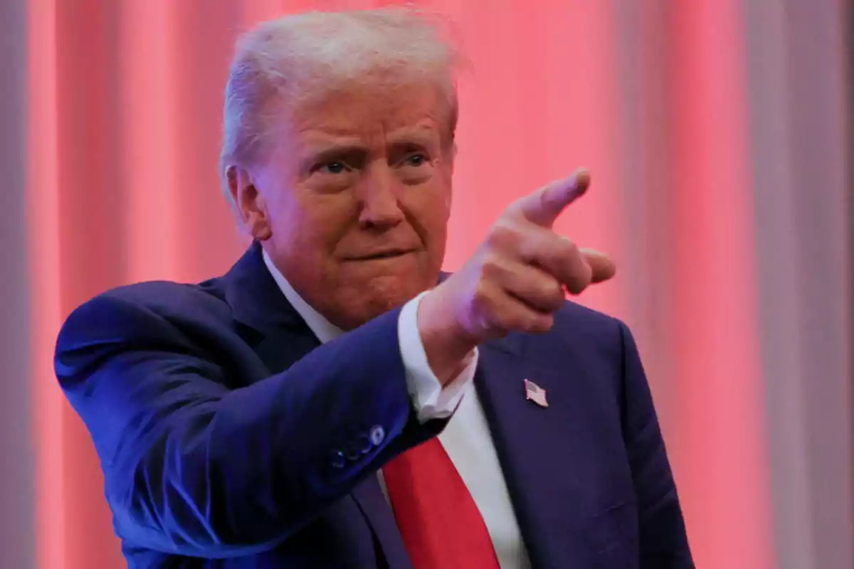 A man in a blue suit and red tie points his finger in an environment with curtains in the background.