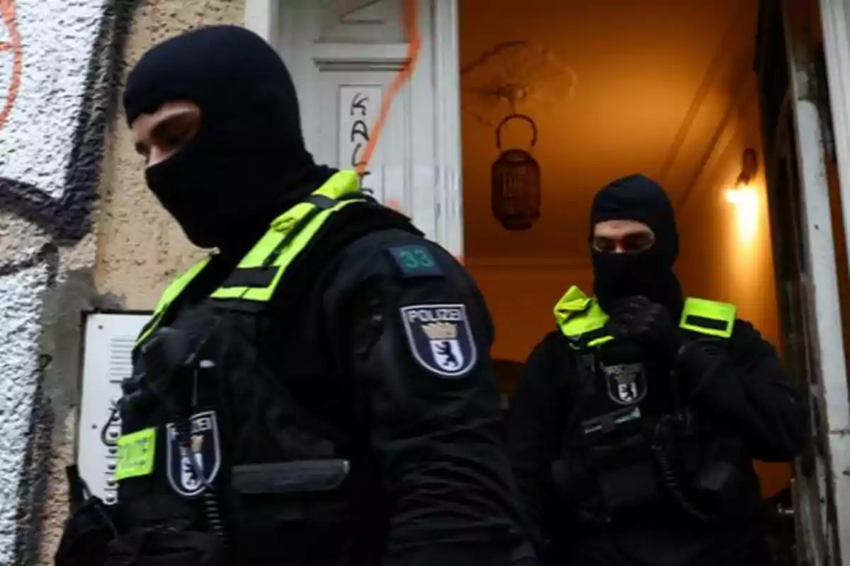 Two police officers wearing balaclavas and high-visibility vests exit a building.