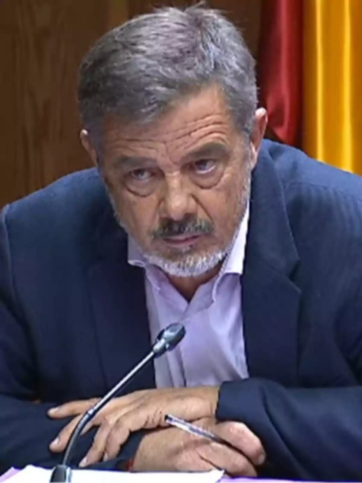 Hombre de cabello canoso y barba sentado frente a un micrófono con expresión seria y fondo de banderas Hombre de cabello canoso y barba sentado frente a un micrófono con expresión seria y fondo de banderas