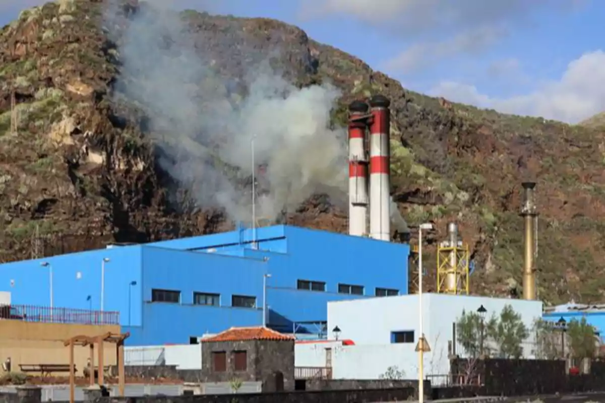 Fábrica de color azul con chimeneas que emiten humo ubicada frente a una montaña Fábrica de color azul con chimeneas que emiten humo ubicada frente a una montaña