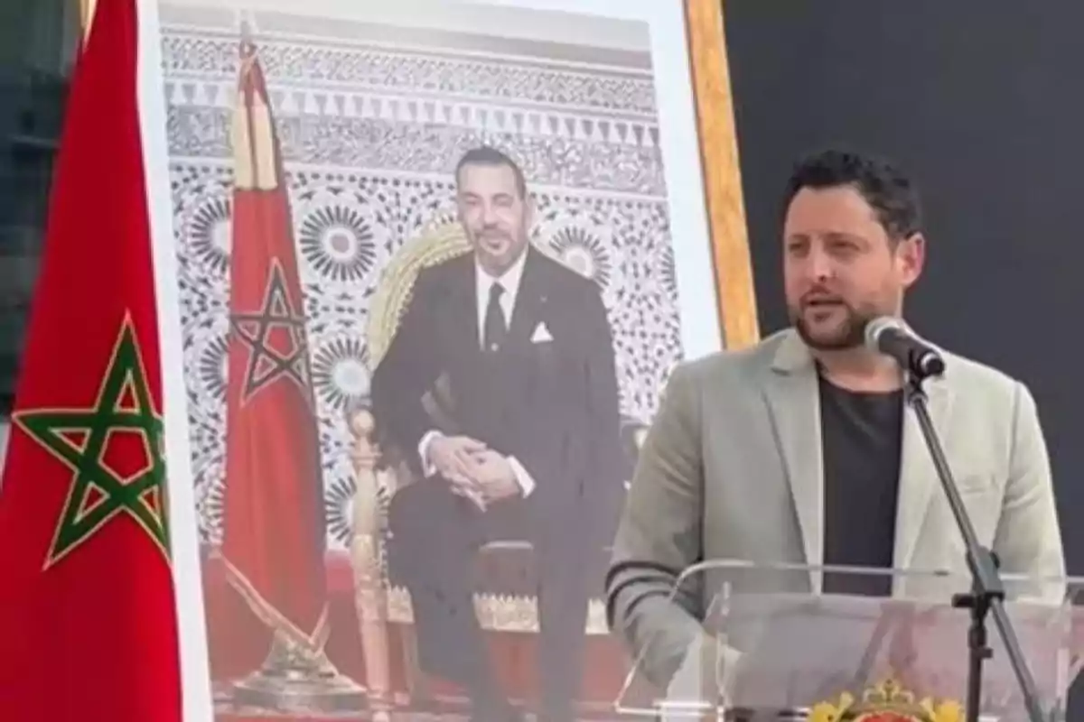 Un hombre de pie junto a un atril con un micrófono, al lado de una bandera de Marruecos y un retrato de una persona sentada en un trono.