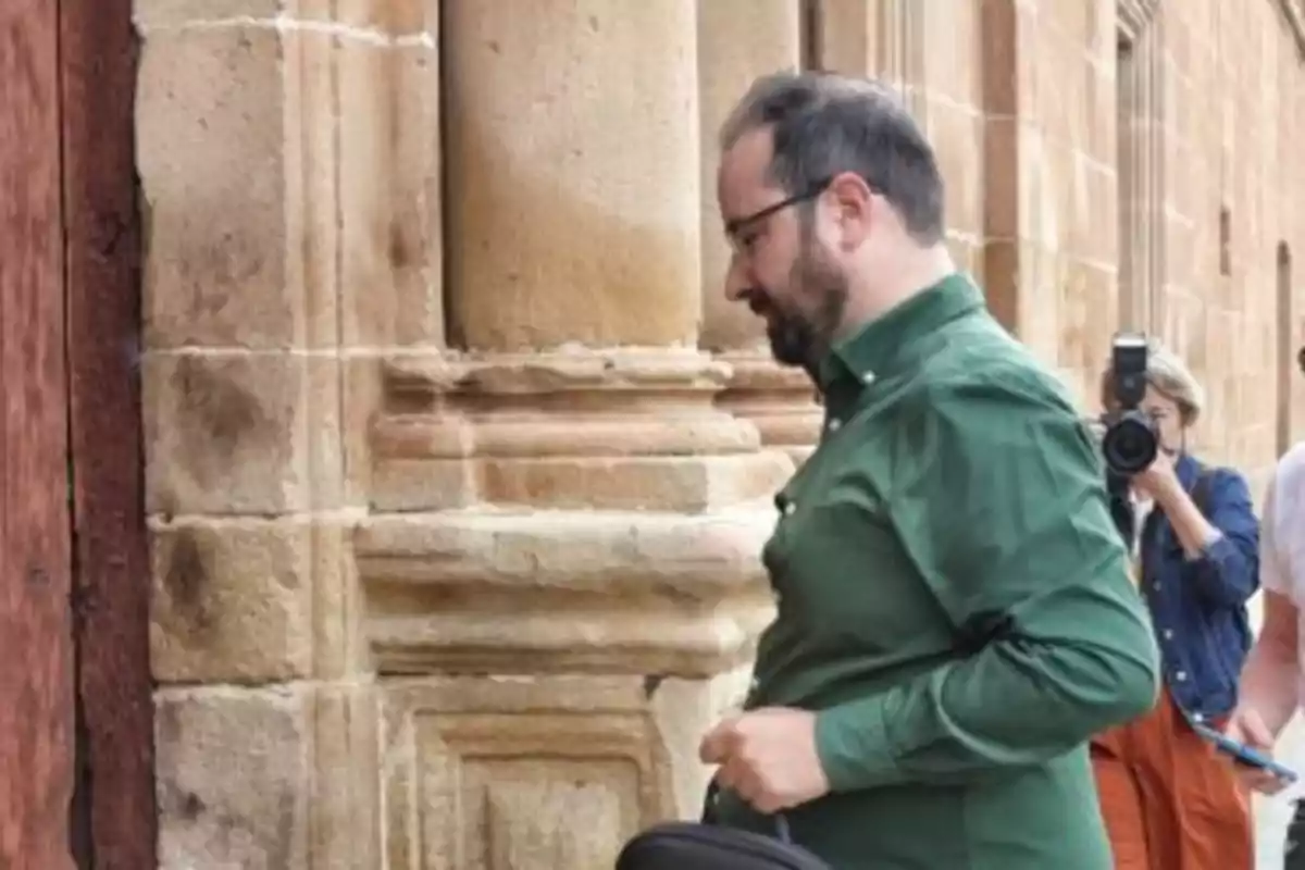 Hombre con camisa verde de perfil caminando junto a una pared de piedra mientras una persona al fondo toma una fotografía Hombre con camisa verde de perfil caminando junto a una pared de piedra mientras una persona al fondo toma una fotografía