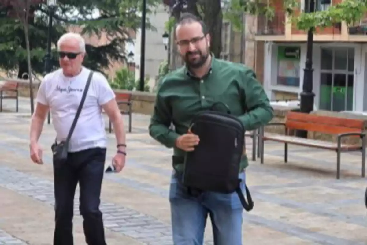 Dos hombres caminando por una plaza, uno lleva una mochila negra y camisa verde, el otro viste camiseta blanca y gafas de sol Dos hombres caminando por una plaza, uno lleva una mochila negra y camisa verde, el otro viste camiseta blanca y gafas de sol