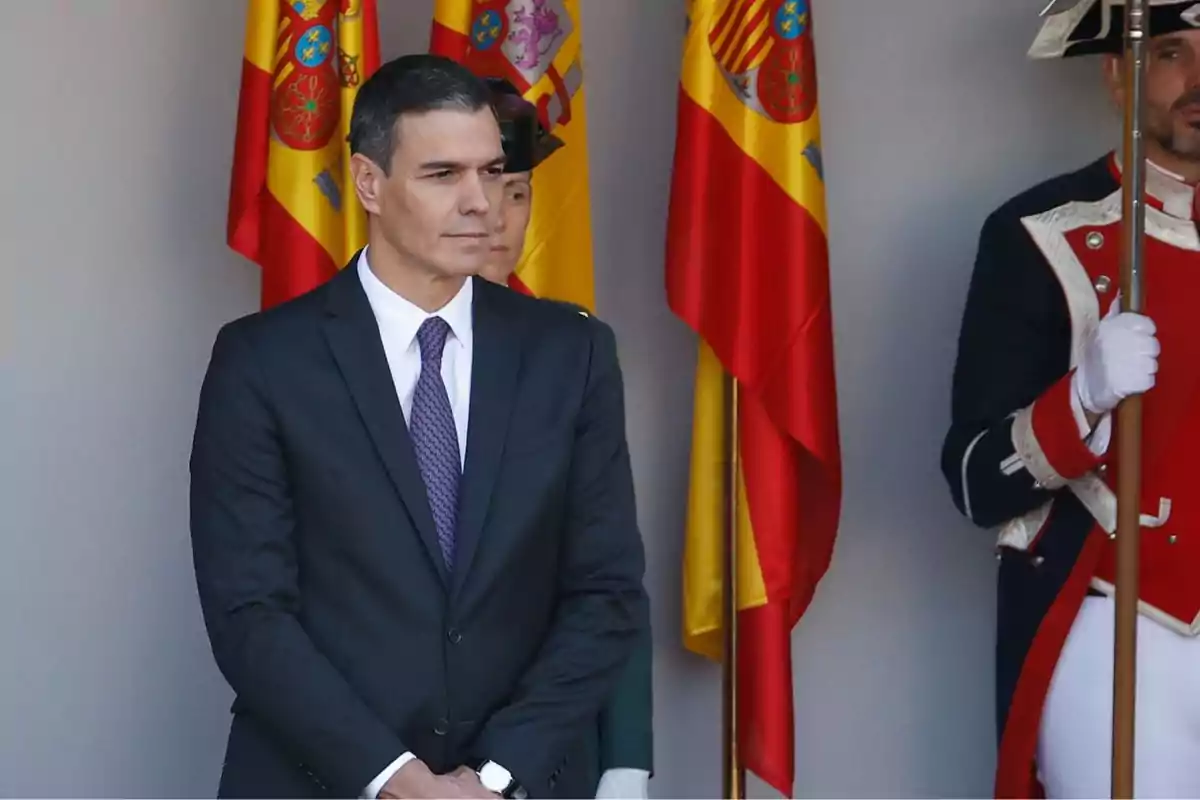 Un hombre de traje oscuro y corbata morada está de pie frente a varias banderas de España, acompañado por un guardia con uniforme ceremonial.