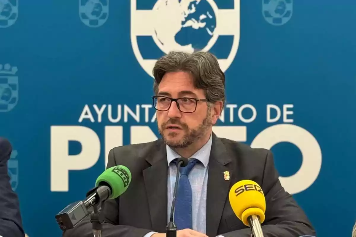 Un hombre con gafas y barba en una conferencia de prensa frente a un fondo azul que dice "Ayuntamiento de Pinto" con micrófonos de colores verde y amarillo.