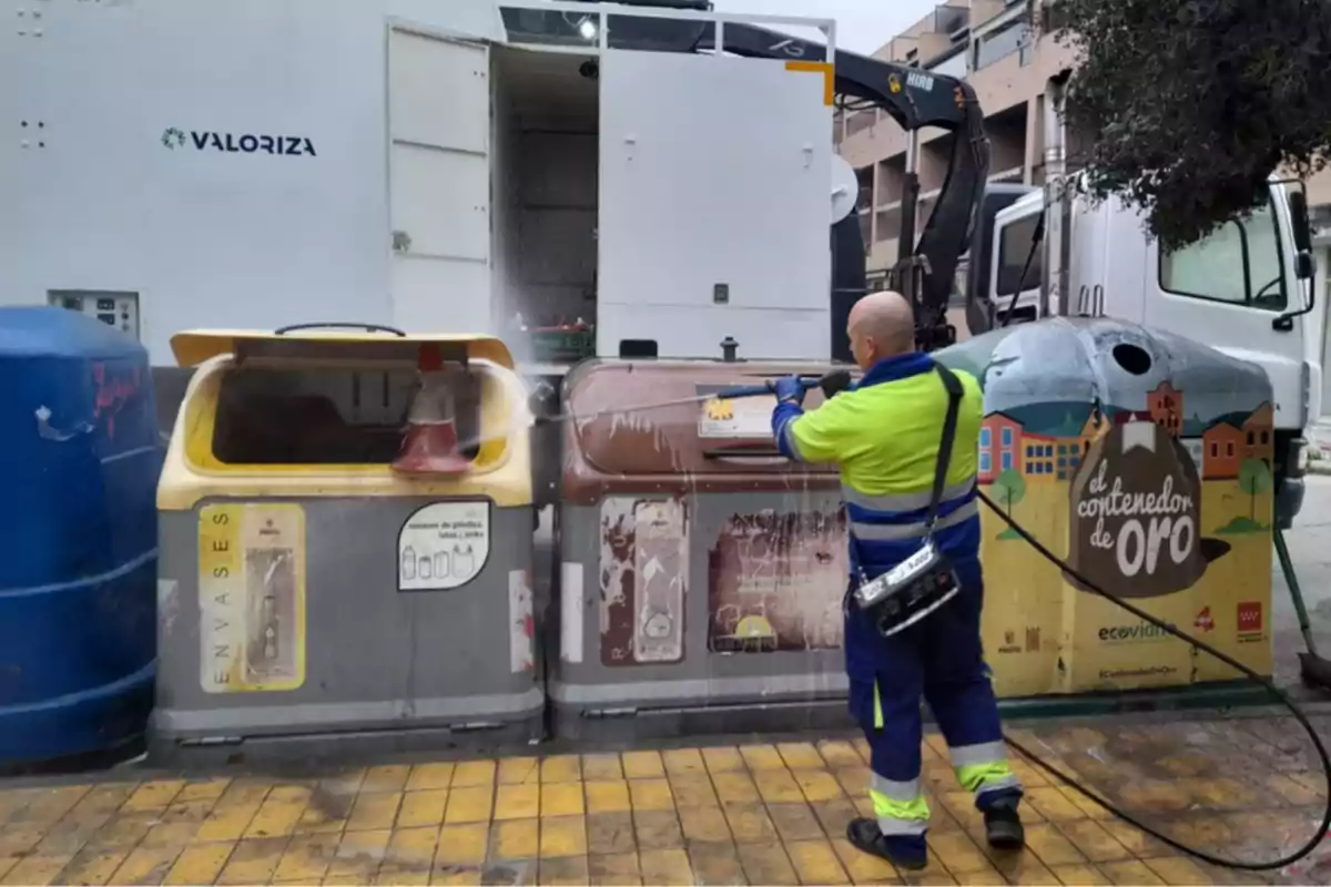 Trabajador de limpieza con uniforme amarillo y azul usando una manguera de alta presión para limpiar contenedores de reciclaje en la calle.