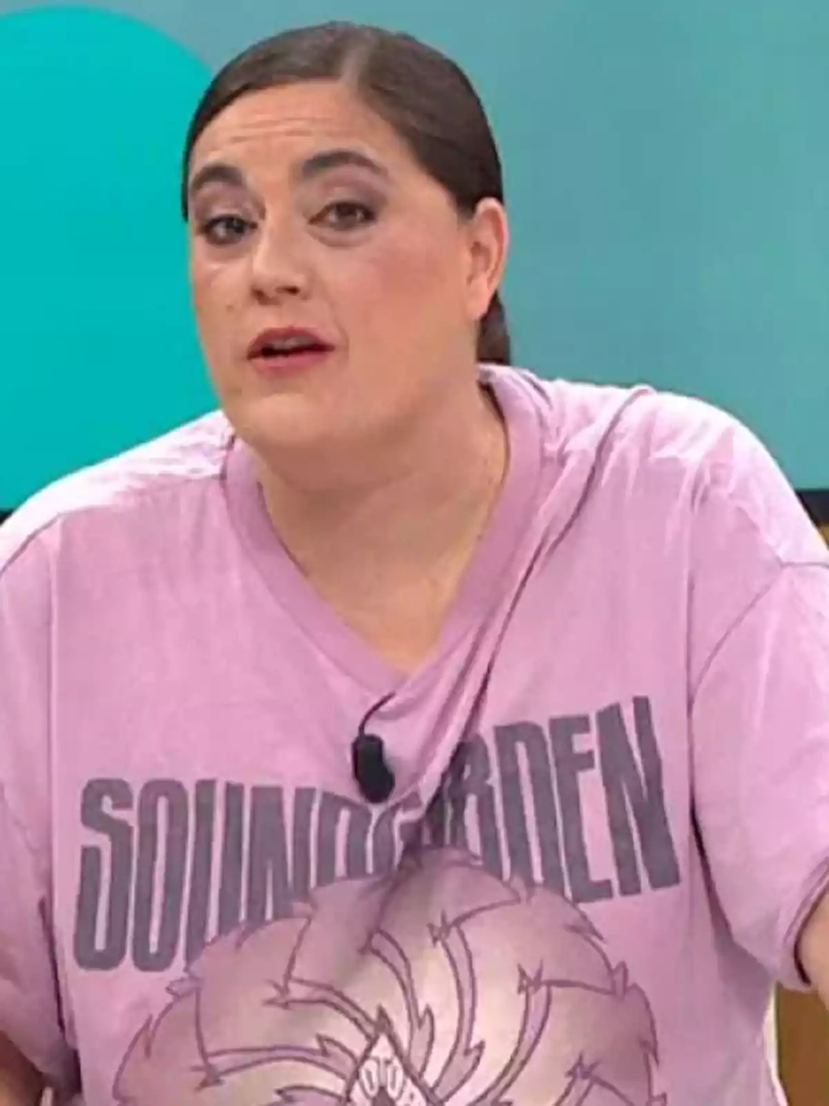 Mujer de cabello oscuro recogido usando una camiseta lila de Soundgarden y un micrófono de solapa, sentada frente a un fondo azul Mujer de cabello oscuro recogido usando una camiseta lila de Soundgarden y un micrófono de solapa, sentada frente a un fondo azul