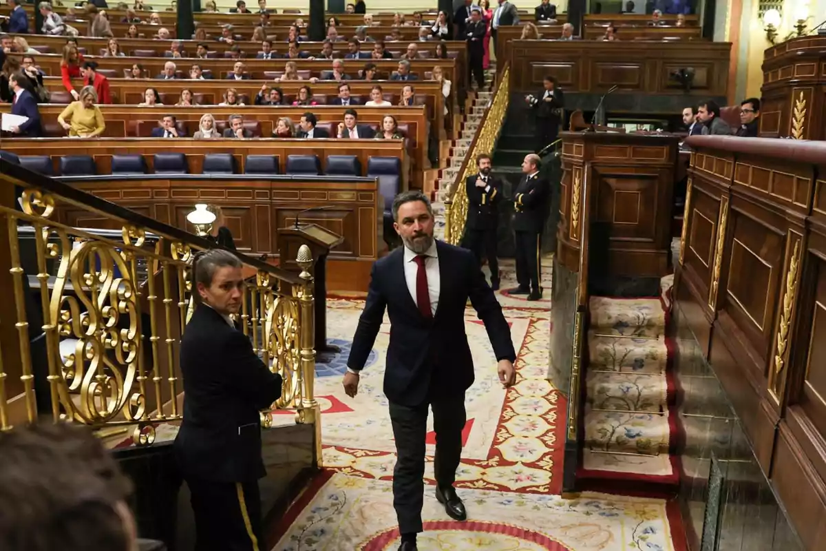 Un hombre con traje oscuro y corbata roja camina por el interior de un parlamento lleno de personas sentadas en sus escaños.