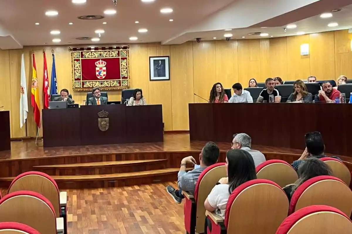 Personas sentadas en un salón de plenos con paredes de madera, varias banderas y un escudo oficial en la pared, mientras algunos asistentes observan desde las butacas. Personas sentadas en un salón de plenos con paredes de madera, varias banderas y un escudo oficial en la pared, mientras algunos asistentes observan desde las butacas.