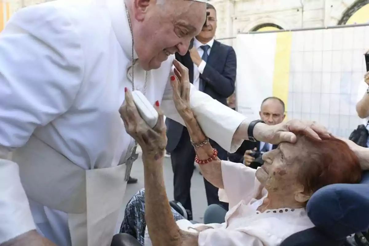 Un hombre vestido de blanco, con una cruz colgando de su cuello, toca la cabeza de una mujer mayor en silla de ruedas mientras ella levanta la mano hacia él.