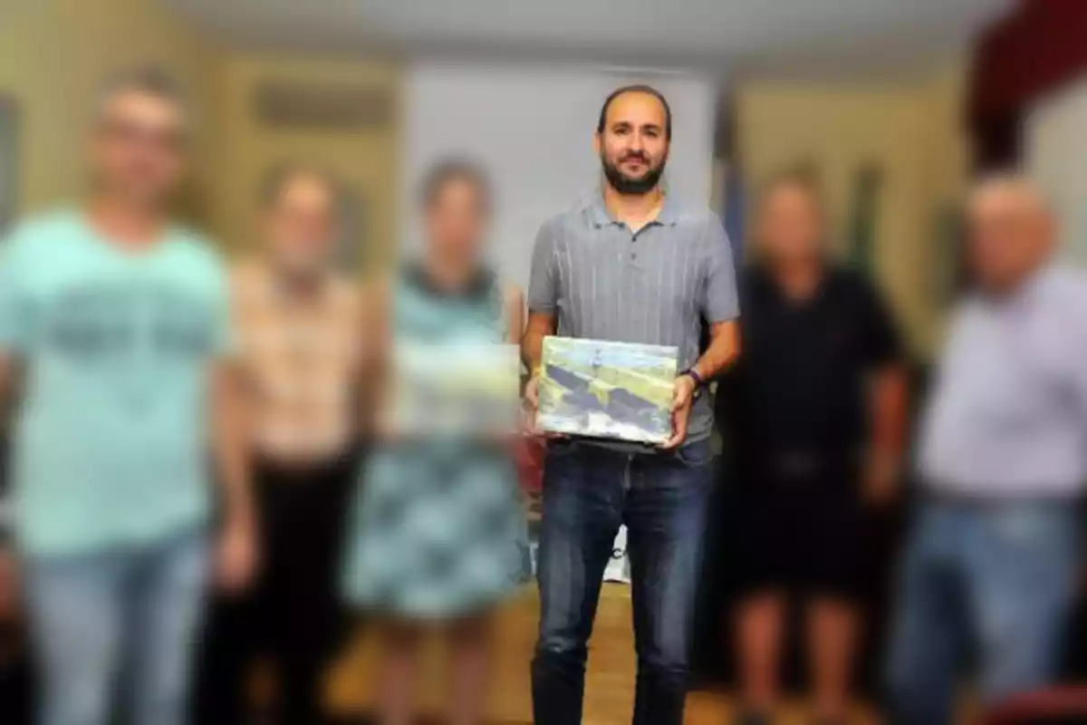 Un hombre de pie al centro sostiene un cuadro mientras varias personas desenfocadas posan a su alrededor en un ambiente interior Un hombre de pie al centro sostiene un cuadro mientras varias personas desenfocadas posan a su alrededor en un ambiente interior
