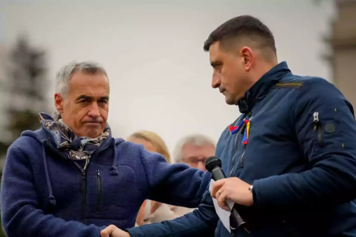 Dos hombres se dan la mano en un evento al aire libre, uno sostiene un micrófono y ambos visten ropa de abrigo.