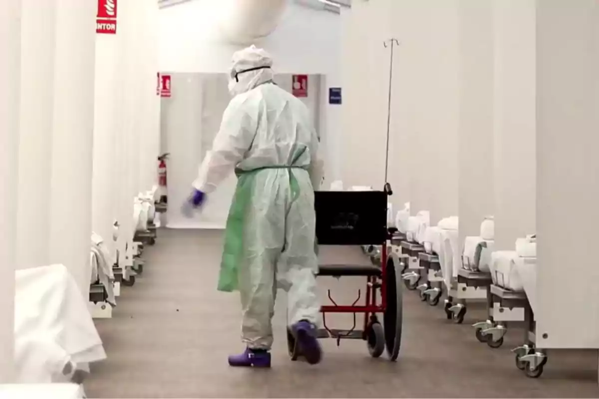 Persona con equipo de protección empujando una silla de ruedas en un pasillo de hospital con camas alineadas a los lados