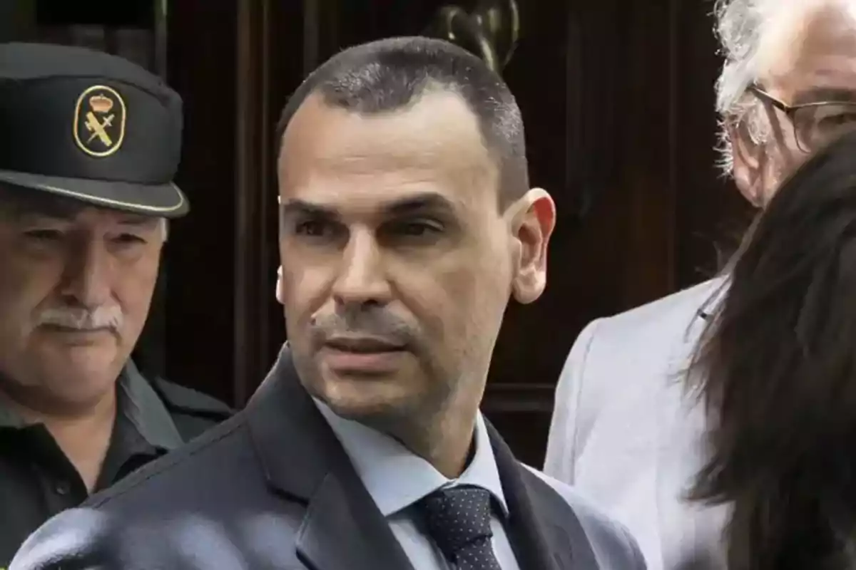 A man in a suit and tie is surrounded by other people, including an officer in uniform and cap. A man in a suit and tie is surrounded by other people, including an officer in uniform and cap.