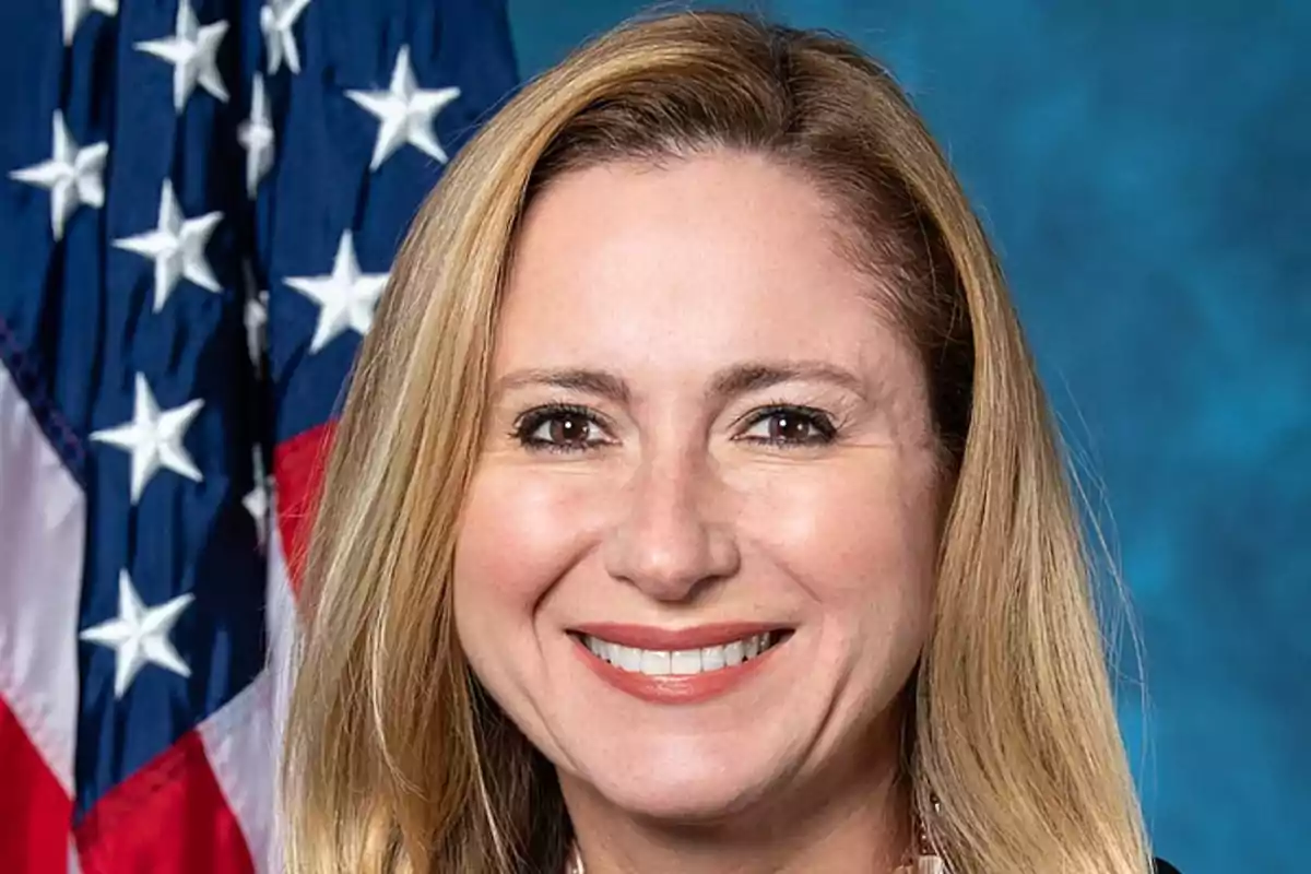 Una mujer sonriente con cabello rubio y una bandera de Estados Unidos en el fondo.