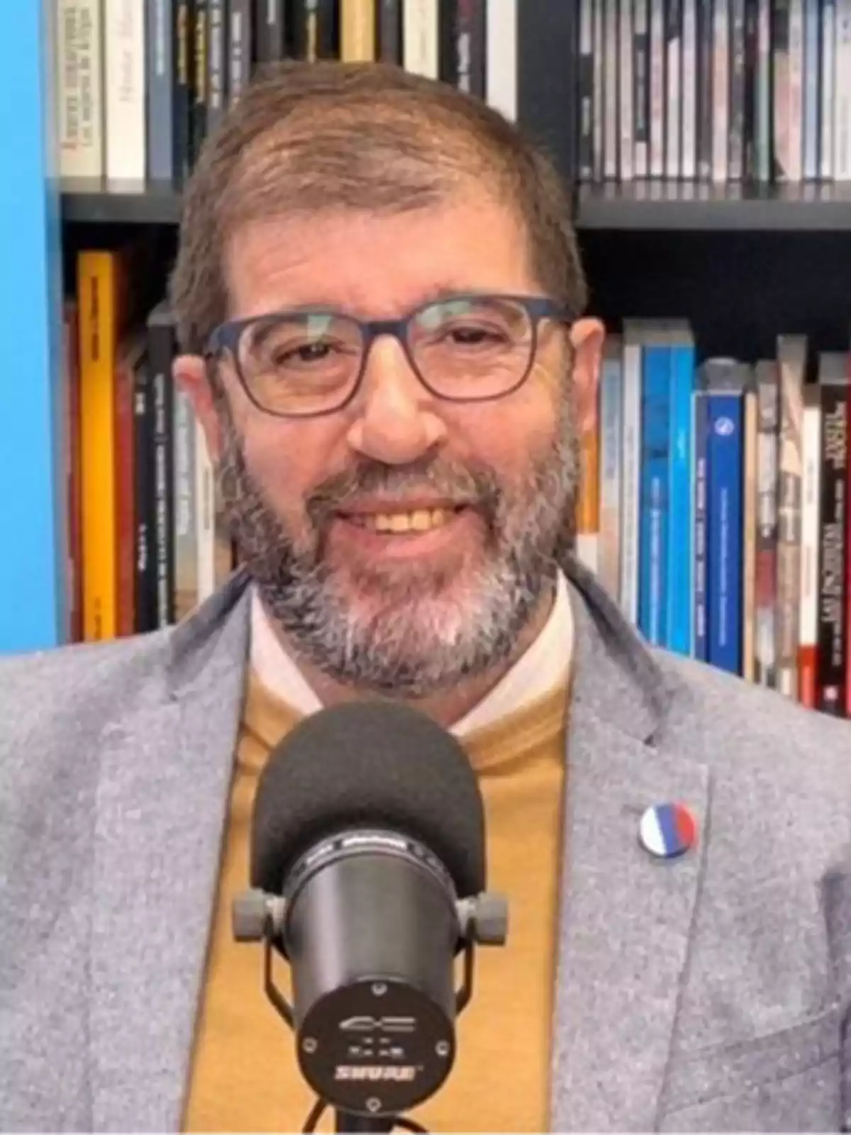 Un hombre con barba y gafas sonríe frente a un micrófono, con una estantería llena de libros detrás. Un hombre con barba y gafas sonríe frente a un micrófono, con una estantería llena de libros detrás.