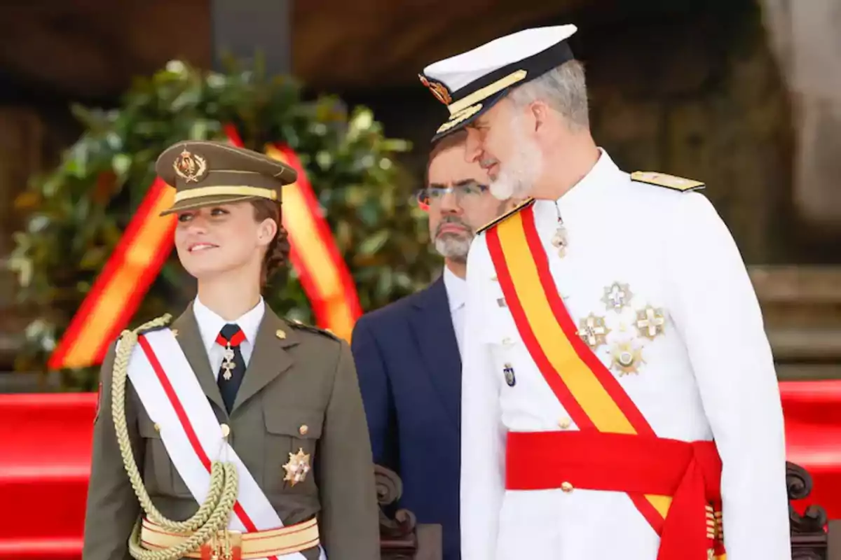 En la imagen se observa a dos personas vestidas con uniformes militares, una de ellas con uniforme verde y la otra con uniforme blanco, ambos con bandas y condecoraciones, mientras que al fondo se aprecia una decoración con cintas rojas y amarillas.