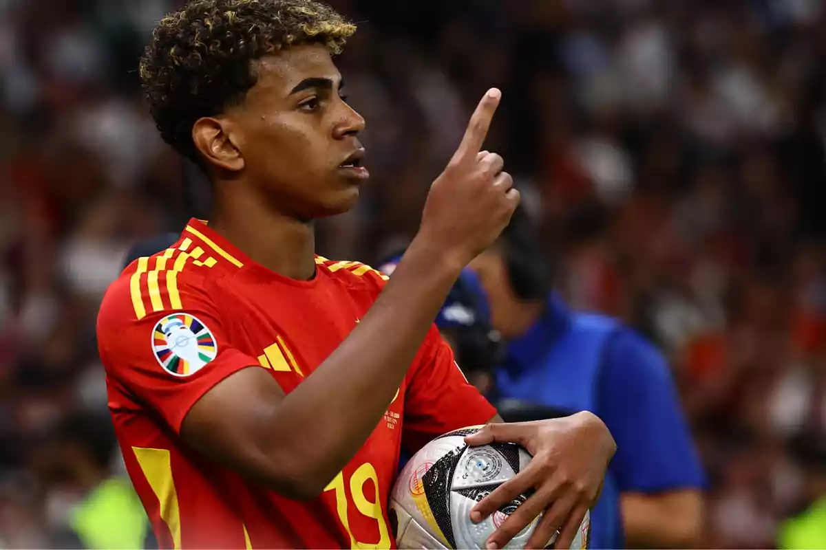 Jugador de fútbol con uniforme rojo y amarillo sosteniendo un balón y señalando con el dedo índice.