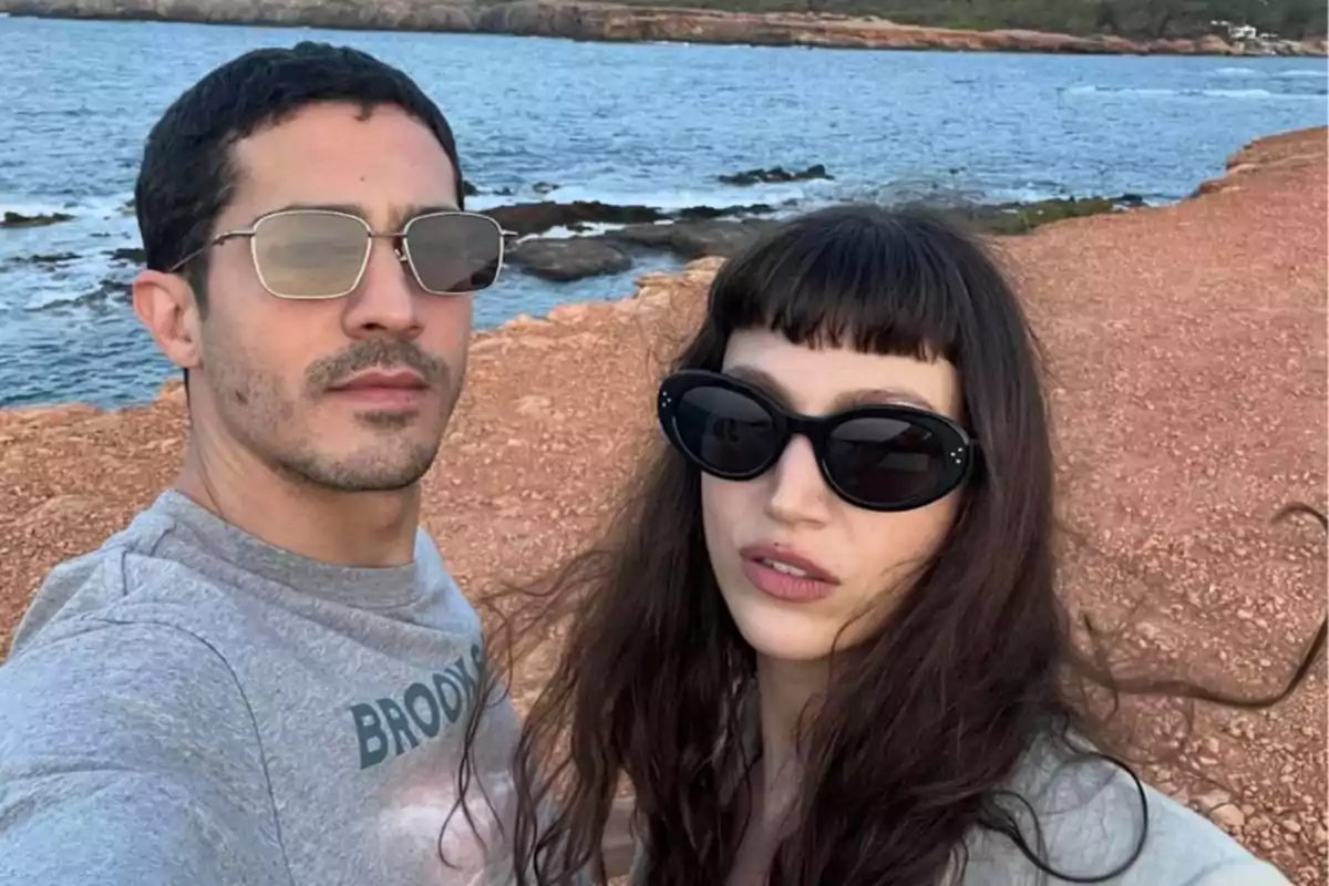 Una pareja con gafas de sol se toma una selfie en un entorno costero con el mar y rocas de fondo.