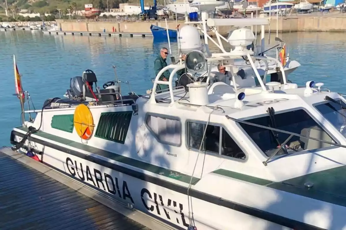 Una embarcación de la Guardia Civil está atracada en un muelle con dos personas a bordo y el agua del puerto reflejando el cielo azul.